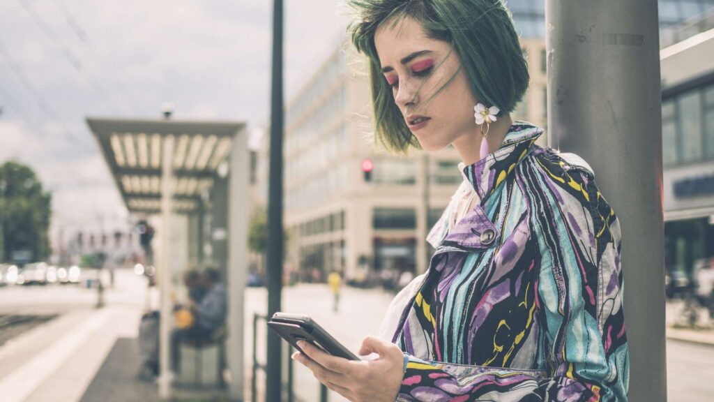 Auffällig bunt gekleidete Frau mit grünen Haaren und pinken Lidschatten schaut auf ein Smartphone an einer Haltestelle